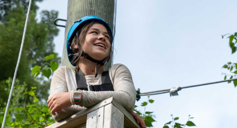A student wearing safety gear smiles as they rest on the platform of a ropes course. 